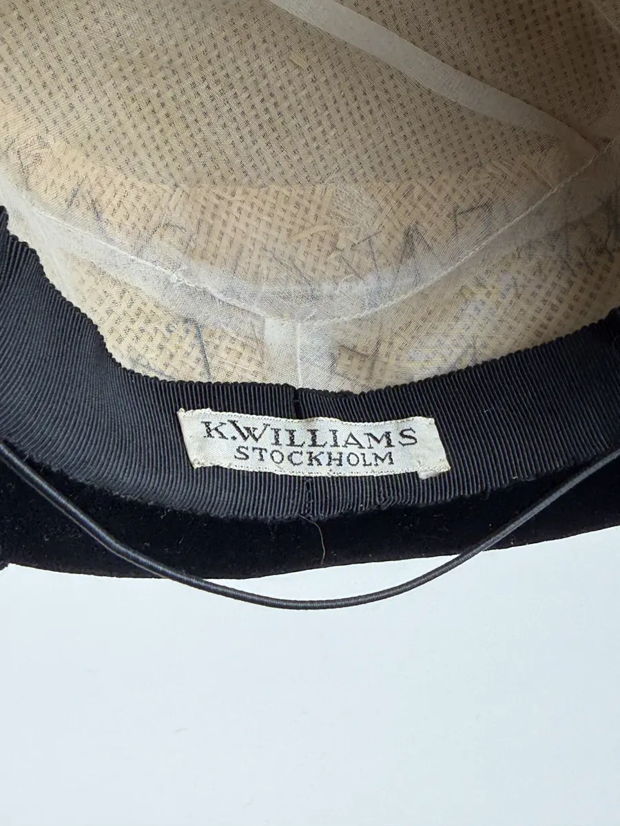 Rund hatt utan brätte. Stomme av styv gles väv av naturfiber klädd med svart sammet. Dekorerad med ett band av en slingrad svart snodd. 