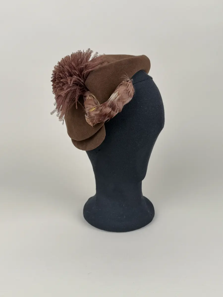 Hatt av asymmetrisk modell utförd i draperade delar av brun filtmaterial. På ena sidan fjäderdekorationer. En boll formad tofs av brunfärgade strutsdun och en fjäderklädd rulle.