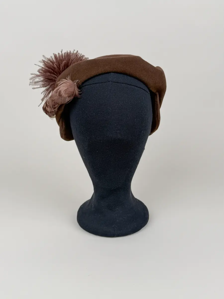 Hatt av asymmetrisk modell utförd i draperade delar av brun filtmaterial. På ena sidan fjäderdekorationer. En boll formad tofs av brunfärgade strutsdun och en fjäderklädd rulle.