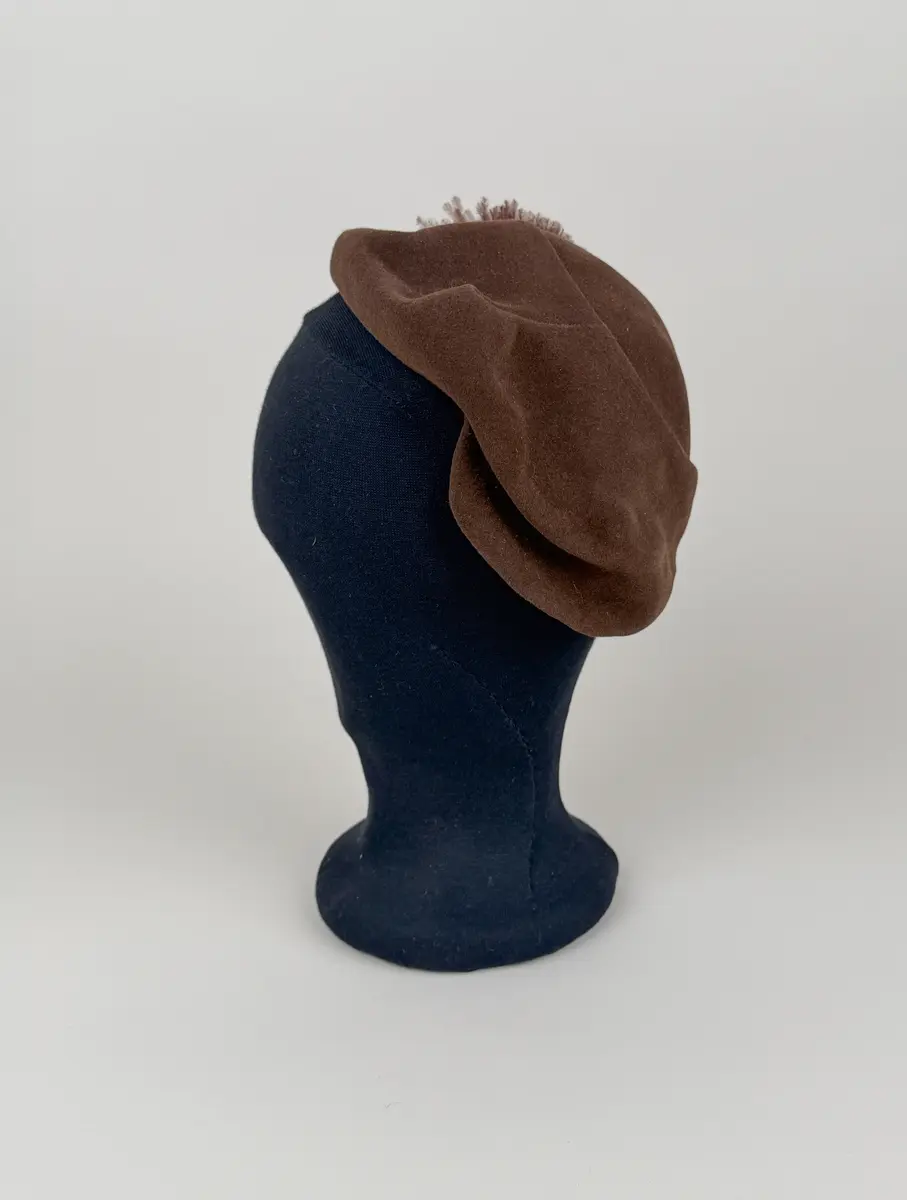 Hatt av asymmetrisk modell utförd i draperade delar av brun filtmaterial. På ena sidan fjäderdekorationer. En boll formad tofs av brunfärgade strutsdun och en fjäderklädd rulle.