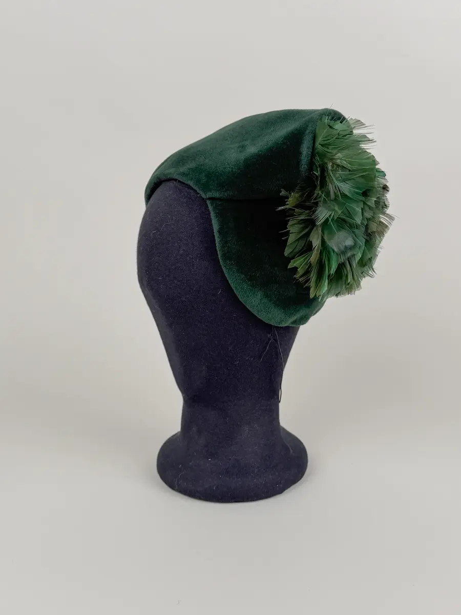 Hat av mörkgrönt ullmaterial med kort sammets-lugg på rätsidan. Formad till en asymmetrisk hätt-liknande form genom veck. Ena sidan slät och andra sidan reser sig i ett trubbigt hörn där en fjäderklädd dekoration är monterad. Kortklippta grönfärgade fjädrar. Innerkanten klädd med svart ripsband.