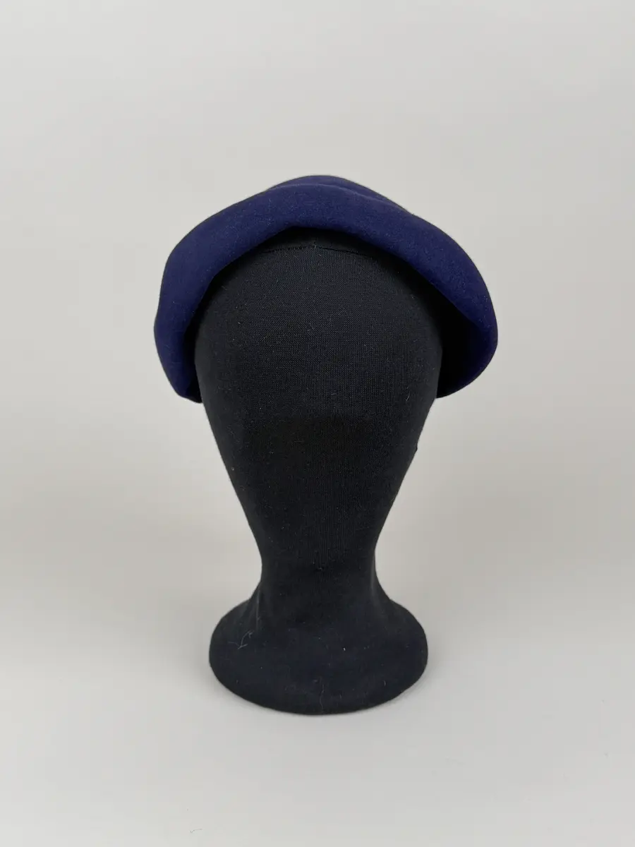 Hatt av moderiktigt modell. Mörkt marinblå. Asymmetrisk toppig konform som lutar åt ena sidan. Från toppen hänger en tofs med dekorativ knut och lång frans. Ripsband längs insidan och en liten kam fastsydd. Etikett med invävt firmanamn.