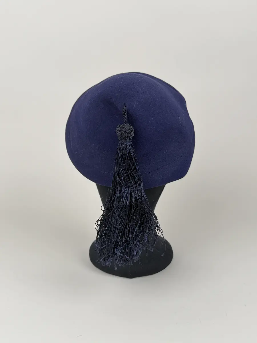 Hatt av moderiktigt modell. Mörkt marinblå. Asymmetrisk toppig konform som lutar åt ena sidan. Från toppen hänger en tofs med dekorativ knut och lång frans. Ripsband längs insidan och en liten kam fastsydd. Etikett med invävt firmanamn.