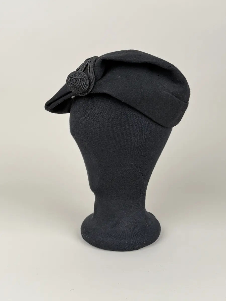 Hatt av moderiktig modell utför i svart ull. Mindre modell, påminner om asymmetrisk stukad basker med ett litet skärm-liknande brätte. Ovanpå hatten en dekoration av i mönster lagda och monterade snoddar som omsluter en snoddklädd rund knopp. Etikett på insidan med vävt firmanamn.