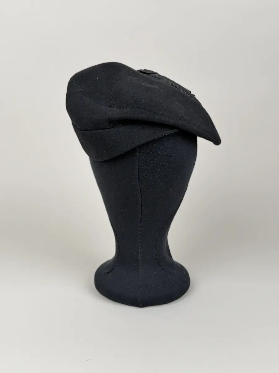 Hatt av moderiktig modell utför i svart ull. Mindre modell, påminner om asymmetrisk stukad basker med ett litet skärm-liknande brätte. Ovanpå hatten en dekoration av i mönster lagda och monterade snoddar som omsluter en snoddklädd rund knopp. Etikett på insidan med vävt firmanamn.