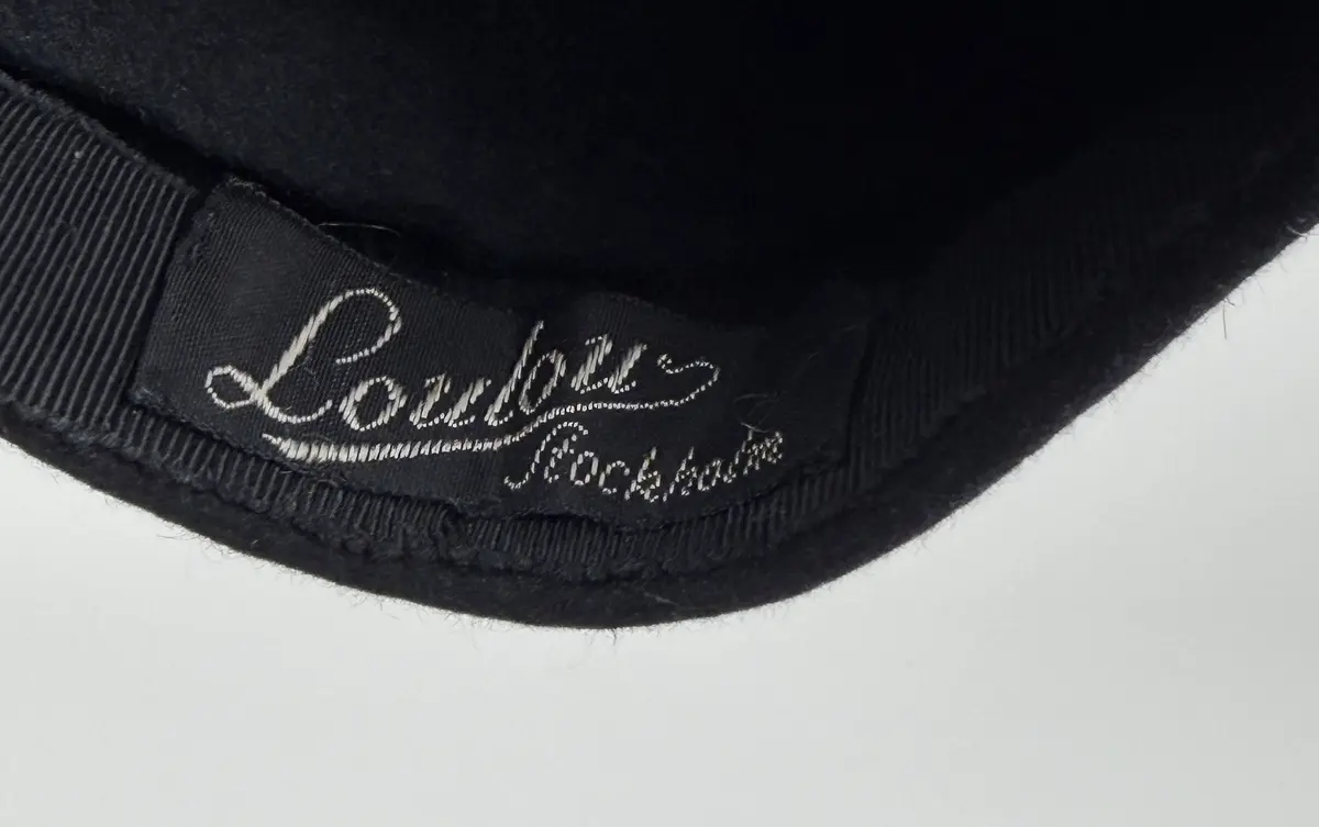 Hatt av moderiktig modell utför i svart ull. Mindre modell, påminner om asymmetrisk stukad basker med ett litet skärm-liknande brätte. Ovanpå hatten en dekoration av i mönster lagda och monterade snoddar som omsluter en snoddklädd rund knopp. Etikett på insidan med vävt firmanamn.