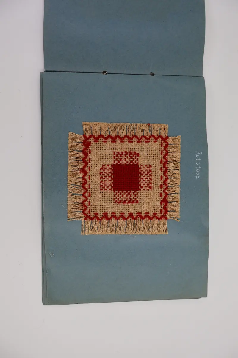 Provbok med stoppsömmar. Egenhändigt tillverkad bok av vikta ark i gråblå kartong sammanfogade med ett slitet rosa sidenband knutet i rosett. Handskriven text i vitt på framsidan: "STOPPNING  IRMA WELIN.".
- På första uppslaget, höger sida står det: "Rutstopp."
Stoppsömmen är sydd med rött ullgarn i form av en kvadrat mitt på den 120 x120 mm stora bottenväven i panamabindning. Fållad med sicksack-bård innanför fransen.
- Nästa uppslag visar prov på två två olika maskstoppar på var sin slätstickad lapp med rätstickad kant längs alla fyra sidor. Lapparna är stickade i oblekt bomullsgarn, maskstopparna i blågrått bomullsgarn i form av en kvadrat mitt på lappen. På den högra sidan står överst: "Äkta maskstopp.".
- Tredje uppslaget, till vänster ytterligare en stickad lapp i oblekt bomullsgarn. Resårstickad med rätstickad kant runt om. Maskstopp i blågrått bomullsgarn i en kvadratisk ruta mitt på lappen.
På höger sida står det med vit text: "Mönsterstopp till gåsögon.". En gles klistrad väv i vitt med mönsterstoppning i rött garn i den ena riktningen och blått garn i den andra riktningen i form av ett gåsöga. Röd broderad bård innanför fransen.
- Fjärde uppslaget, till höger; ett prov på beige gabardintyg, mönsterstoppat/konststoppat på två ställen med tråd i samma färg. Text högst upp: "Mönsterstopp-till-gabardin.".
- Nästa uppslag, till höger visar ett rutigt kostym-/dräkttyg vävt i kypert med prov på en fin lagning. Överst står det: "Lagning-av-revor.".
- Sista uppslaget, till höger visar två prover på tyllstoppning. En kvadratisk kartongbit 70 x 70 mm med två nyanser rött garn trätt upp och ned igenom kartongen till ett nät. Trädning/stopp med blått garn över halva ytan. Intill sitter en bit vit tyll med en lagning/stopp mitt på. Text överst på sidan: "Tyllstopp.".