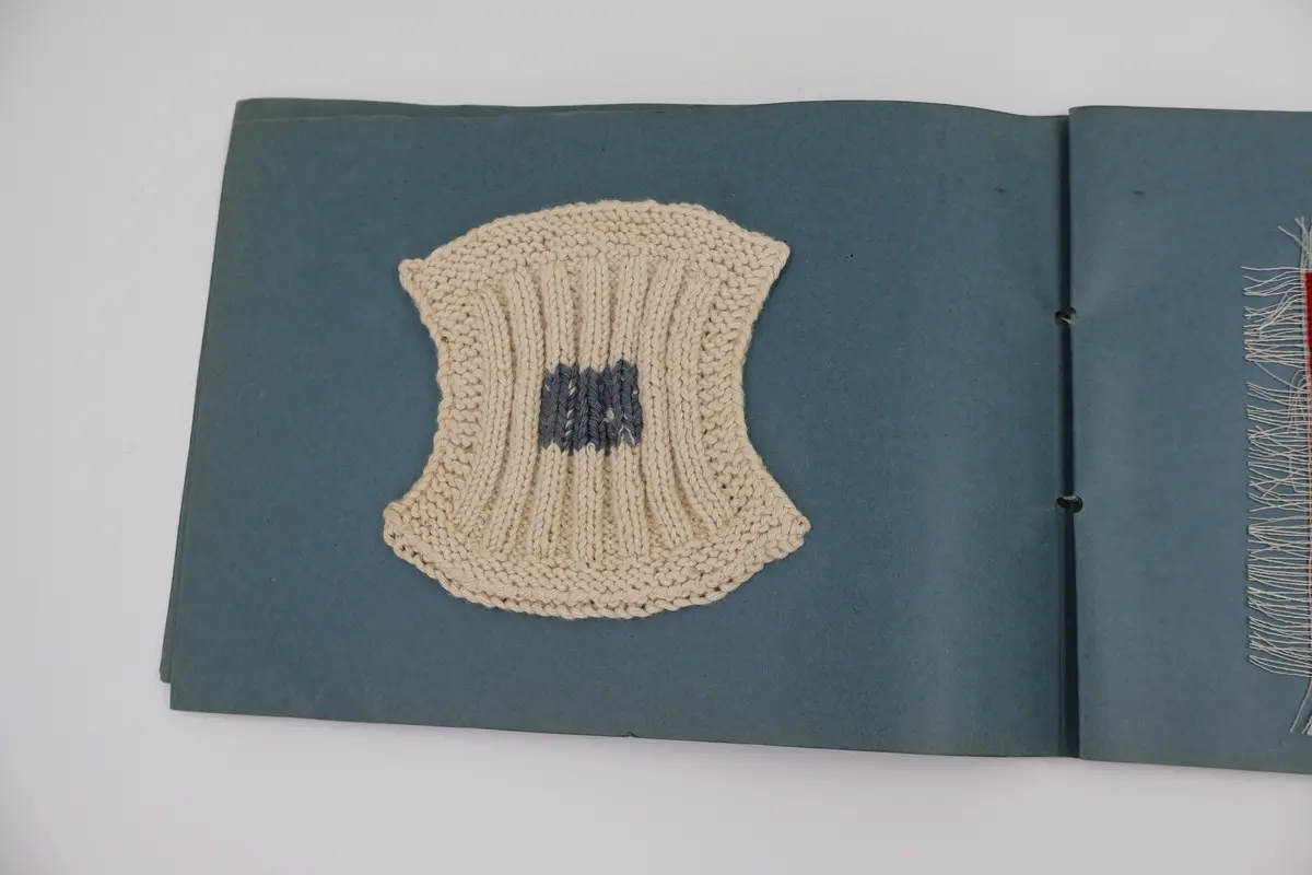 Provbok med stoppsömmar. Egenhändigt tillverkad bok av vikta ark i gråblå kartong sammanfogade med ett slitet rosa sidenband knutet i rosett. Handskriven text i vitt på framsidan: "STOPPNING  IRMA WELIN.".
- På första uppslaget, höger sida står det: "Rutstopp."
Stoppsömmen är sydd med rött ullgarn i form av en kvadrat mitt på den 120 x120 mm stora bottenväven i panamabindning. Fållad med sicksack-bård innanför fransen.
- Nästa uppslag visar prov på två två olika maskstoppar på var sin slätstickad lapp med rätstickad kant längs alla fyra sidor. Lapparna är stickade i oblekt bomullsgarn, maskstopparna i blågrått bomullsgarn i form av en kvadrat mitt på lappen. På den högra sidan står överst: "Äkta maskstopp.".
- Tredje uppslaget, till vänster ytterligare en stickad lapp i oblekt bomullsgarn. Resårstickad med rätstickad kant runt om. Maskstopp i blågrått bomullsgarn i en kvadratisk ruta mitt på lappen.
På höger sida står det med vit text: "Mönsterstopp till gåsögon.". En gles klistrad väv i vitt med mönsterstoppning i rött garn i den ena riktningen och blått garn i den andra riktningen i form av ett gåsöga. Röd broderad bård innanför fransen.
- Fjärde uppslaget, till höger; ett prov på beige gabardintyg, mönsterstoppat/konststoppat på två ställen med tråd i samma färg. Text högst upp: "Mönsterstopp-till-gabardin.".
- Nästa uppslag, till höger visar ett rutigt kostym-/dräkttyg vävt i kypert med prov på en fin lagning. Överst står det: "Lagning-av-revor.".
- Sista uppslaget, till höger visar två prover på tyllstoppning. En kvadratisk kartongbit 70 x 70 mm med två nyanser rött garn trätt upp och ned igenom kartongen till ett nät. Trädning/stopp med blått garn över halva ytan. Intill sitter en bit vit tyll med en lagning/stopp mitt på. Text överst på sidan: "Tyllstopp.".