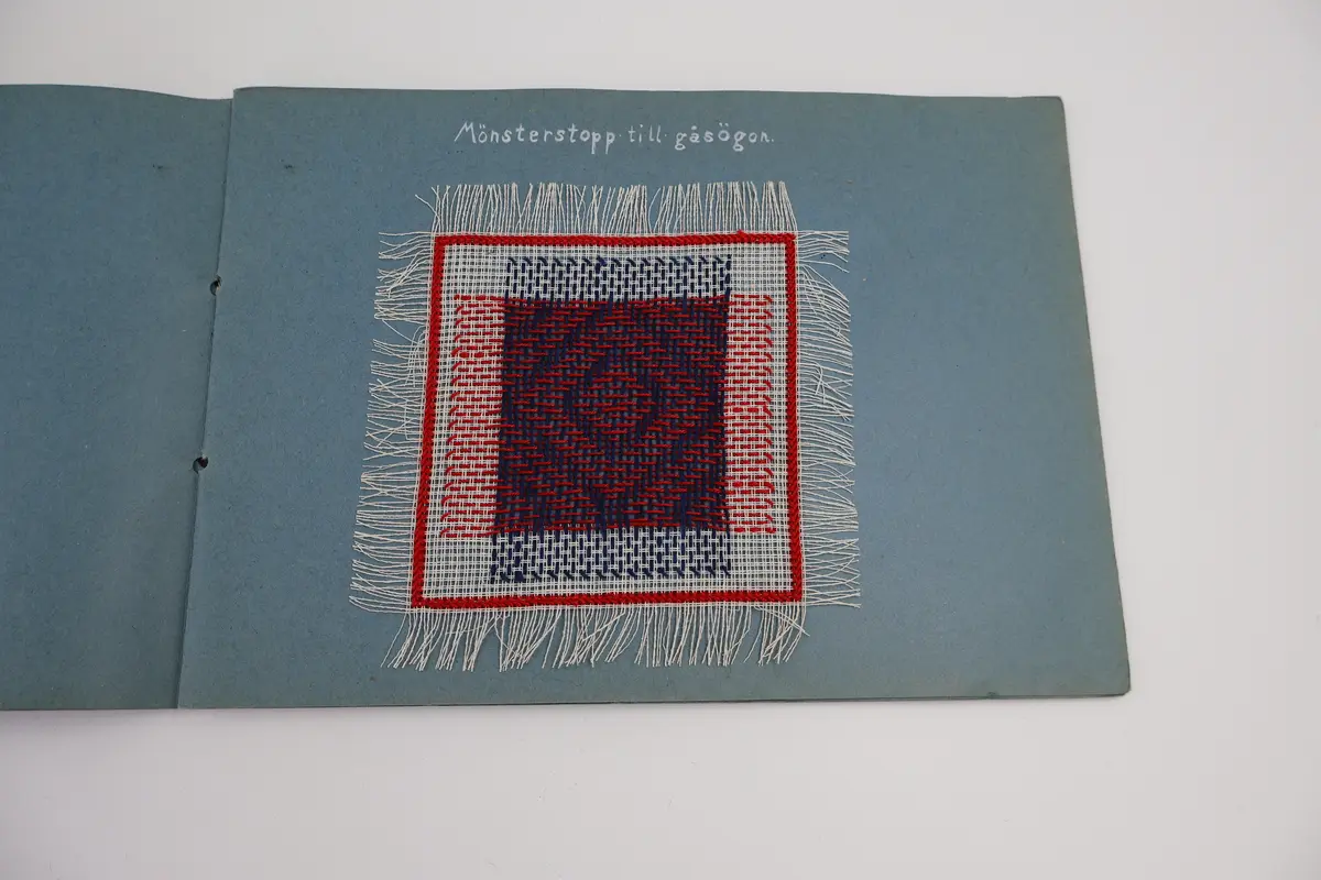 Provbok med stoppsömmar. Egenhändigt tillverkad bok av vikta ark i gråblå kartong sammanfogade med ett slitet rosa sidenband knutet i rosett. Handskriven text i vitt på framsidan: "STOPPNING  IRMA WELIN.".
- På första uppslaget, höger sida står det: "Rutstopp."
Stoppsömmen är sydd med rött ullgarn i form av en kvadrat mitt på den 120 x120 mm stora bottenväven i panamabindning. Fållad med sicksack-bård innanför fransen.
- Nästa uppslag visar prov på två två olika maskstoppar på var sin slätstickad lapp med rätstickad kant längs alla fyra sidor. Lapparna är stickade i oblekt bomullsgarn, maskstopparna i blågrått bomullsgarn i form av en kvadrat mitt på lappen. På den högra sidan står överst: "Äkta maskstopp.".
- Tredje uppslaget, till vänster ytterligare en stickad lapp i oblekt bomullsgarn. Resårstickad med rätstickad kant runt om. Maskstopp i blågrått bomullsgarn i en kvadratisk ruta mitt på lappen.
På höger sida står det med vit text: "Mönsterstopp till gåsögon.". En gles klistrad väv i vitt med mönsterstoppning i rött garn i den ena riktningen och blått garn i den andra riktningen i form av ett gåsöga. Röd broderad bård innanför fransen.
- Fjärde uppslaget, till höger; ett prov på beige gabardintyg, mönsterstoppat/konststoppat på två ställen med tråd i samma färg. Text högst upp: "Mönsterstopp-till-gabardin.".
- Nästa uppslag, till höger visar ett rutigt kostym-/dräkttyg vävt i kypert med prov på en fin lagning. Överst står det: "Lagning-av-revor.".
- Sista uppslaget, till höger visar två prover på tyllstoppning. En kvadratisk kartongbit 70 x 70 mm med två nyanser rött garn trätt upp och ned igenom kartongen till ett nät. Trädning/stopp med blått garn över halva ytan. Intill sitter en bit vit tyll med en lagning/stopp mitt på. Text överst på sidan: "Tyllstopp.".