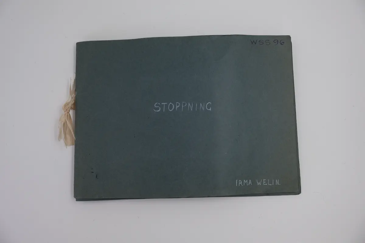 Provbok med stoppsömmar. Egenhändigt tillverkad bok av vikta ark i gråblå kartong sammanfogade med ett slitet rosa sidenband knutet i rosett. Handskriven text i vitt på framsidan: "STOPPNING  IRMA WELIN.".
- På första uppslaget, höger sida står det: "Rutstopp."
Stoppsömmen är sydd med rött ullgarn i form av en kvadrat mitt på den 120 x120 mm stora bottenväven i panamabindning. Fållad med sicksack-bård innanför fransen.
- Nästa uppslag visar prov på två två olika maskstoppar på var sin slätstickad lapp med rätstickad kant längs alla fyra sidor. Lapparna är stickade i oblekt bomullsgarn, maskstopparna i blågrått bomullsgarn i form av en kvadrat mitt på lappen. På den högra sidan står överst: "Äkta maskstopp.".
- Tredje uppslaget, till vänster ytterligare en stickad lapp i oblekt bomullsgarn. Resårstickad med rätstickad kant runt om. Maskstopp i blågrått bomullsgarn i en kvadratisk ruta mitt på lappen.
På höger sida står det med vit text: "Mönsterstopp till gåsögon.". En gles klistrad väv i vitt med mönsterstoppning i rött garn i den ena riktningen och blått garn i den andra riktningen i form av ett gåsöga. Röd broderad bård innanför fransen.
- Fjärde uppslaget, till höger; ett prov på beige gabardintyg, mönsterstoppat/konststoppat på två ställen med tråd i samma färg. Text högst upp: "Mönsterstopp-till-gabardin.".
- Nästa uppslag, till höger visar ett rutigt kostym-/dräkttyg vävt i kypert med prov på en fin lagning. Överst står det: "Lagning-av-revor.".
- Sista uppslaget, till höger visar två prover på tyllstoppning. En kvadratisk kartongbit 70 x 70 mm med två nyanser rött garn trätt upp och ned igenom kartongen till ett nät. Trädning/stopp med blått garn över halva ytan. Intill sitter en bit vit tyll med en lagning/stopp mitt på. Text överst på sidan: "Tyllstopp.".