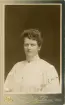 Fröken Clara Schander i höghalsad vit blus med broderier (s.k. engelskt broderi), Runt halsen syns en tunn klockkedja och en brosch.
Bröstbild, halvprofil. Ateljéfoto.
I nedre högra hörnet en autograf: 