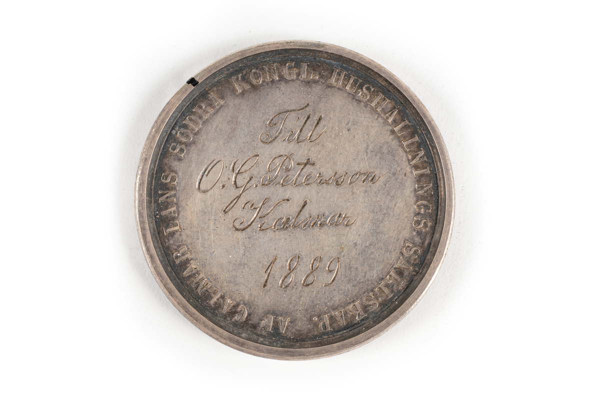 Medalj av silver utdelad av Kalmar läns hushållningssällskap, sannolikt som pris eller belöning för arbete, skicklighet eller prestation inom lantbruk eller liknande.