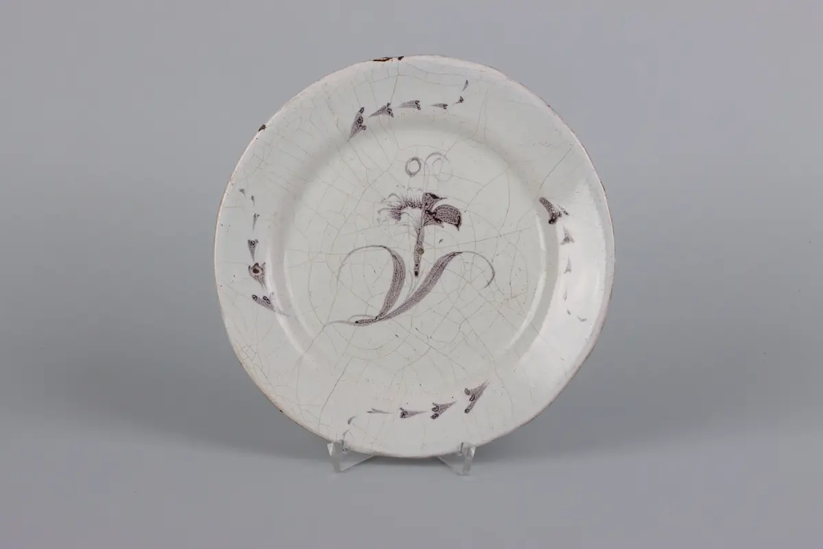  Fat, runt med slätt brätte av vitglaserad fajans. Målad dekor i lila med nejlika i spegeln och fyra rankor på brättet. Oglaserad fotring, på baksidan märken efter sättstöd. Osignerat. Glasyr krackelerad.