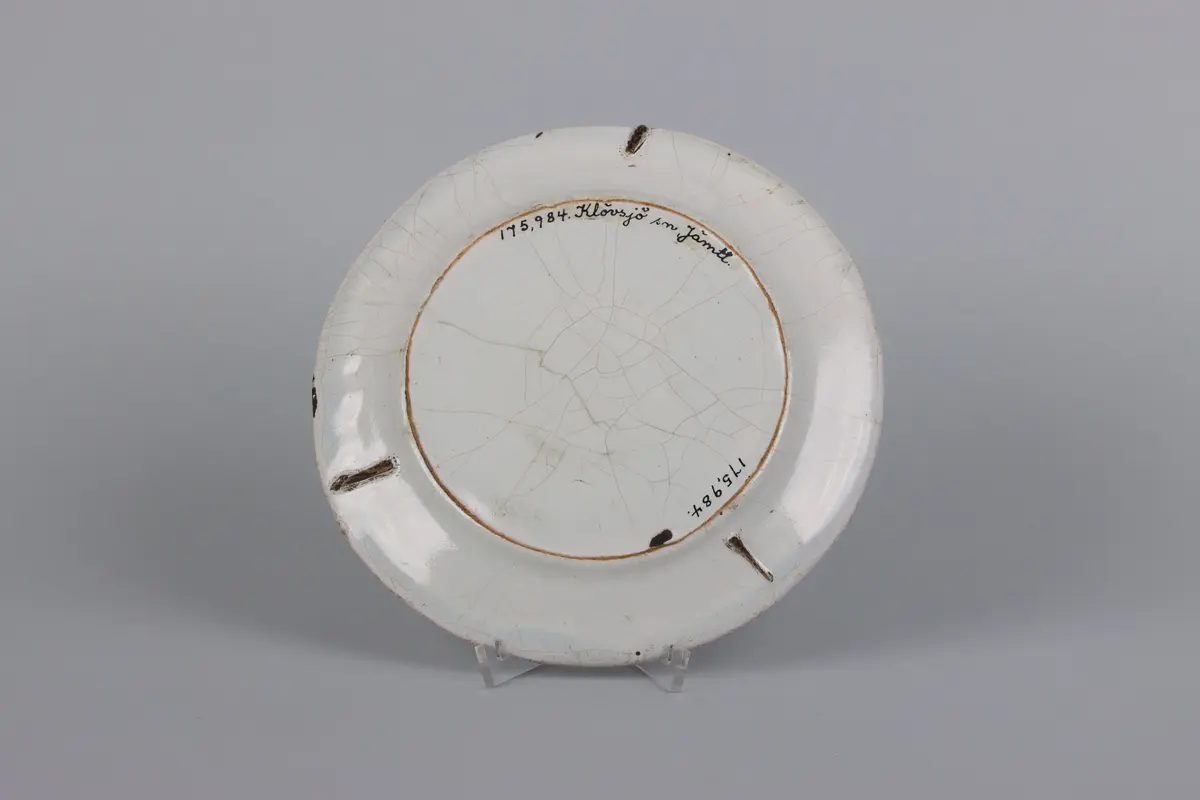  Fat, runt med slätt brätte av vitglaserad fajans. Målad dekor i lila med nejlika i spegeln och fyra rankor på brättet. Oglaserad fotring, på baksidan märken efter sättstöd. Osignerat. Glasyr krackelerad.