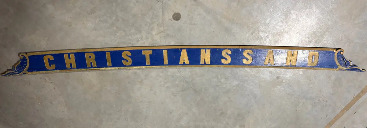Skilt, angivelig fra et skip. Utført i tre, en lang planke, som er utskåret i begge ender. Risset inn ramme og dekor, samt skrift "Christianssand". All dekor er utført i gull, på blå bakgrunn. Umalt på baksiden. 