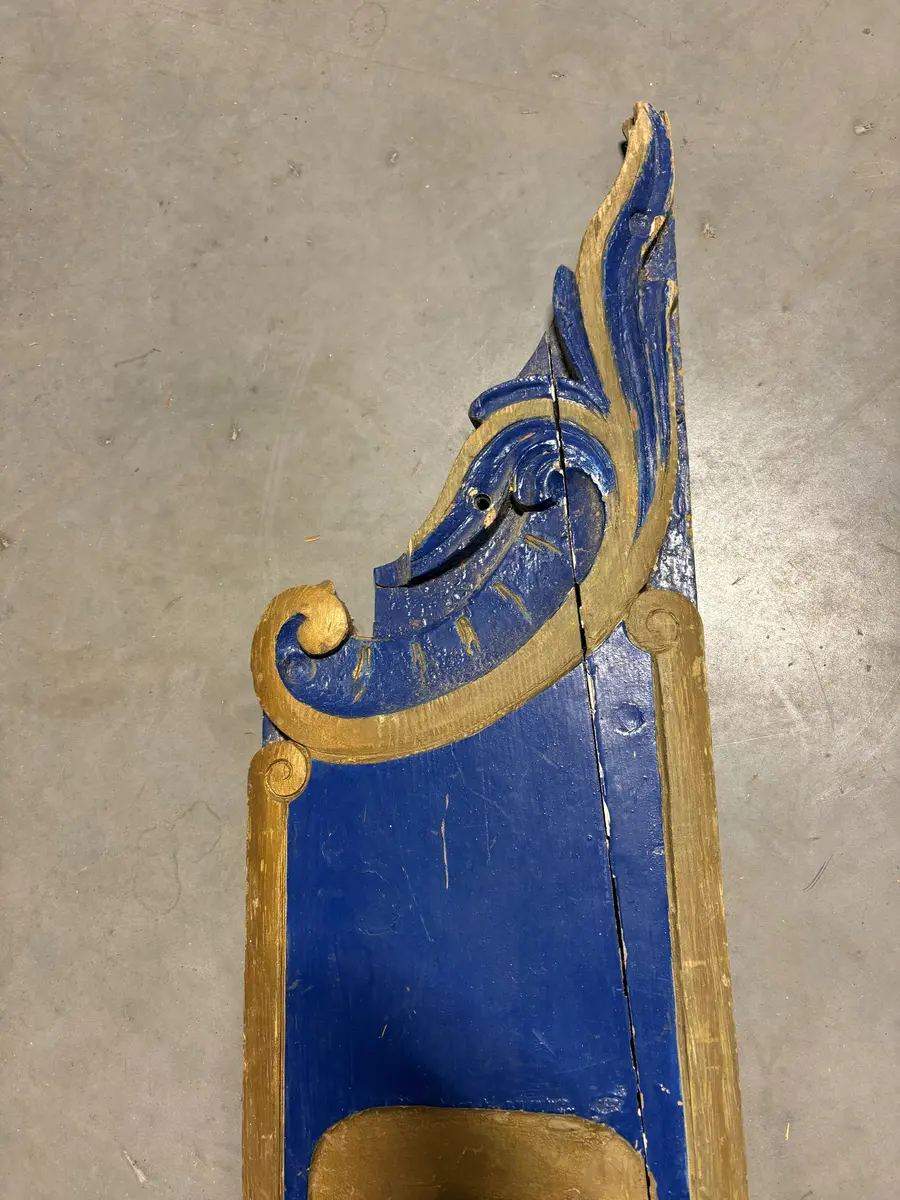 Skilt, angivelig fra et skip. Utført i tre, en lang planke, som er utskåret i begge ender. Risset inn ramme og dekor, samt skrift "Christianssand". All dekor er utført i gull, på blå bakgrunn. Umalt på baksiden. 