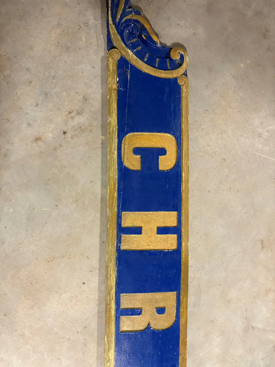 Skilt, angivelig fra et skip. Utført i tre, en lang planke, som er utskåret i begge ender. Risset inn ramme og dekor, samt skrift "Christianssand". All dekor er utført i gull, på blå bakgrunn. Umalt på baksiden. 