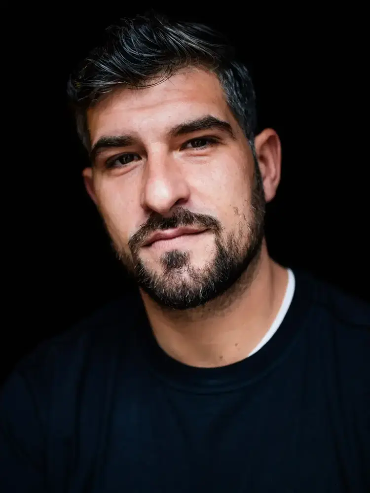 Portrettfotografi i farger av Leo Ajkic. Mann med svart genser, mørke øyne og mørkt hår med noen gråe striper. Han har skjegg og bart.