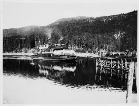 D/F Rjukanfoss (etter ombygging) på vei inn til Tinnoset. Åpne kassevogner Litra L4 med pressenning ombord. To menn stående på brygga.