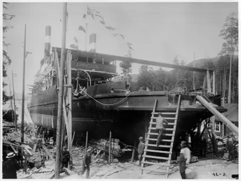 D/F Ammonia på slippen på Tinnoset, mann på vei opp trestige opp på baugen. Flagg henger fra baugen og opp i flaggstanga. Mennesker stående rundt båten.