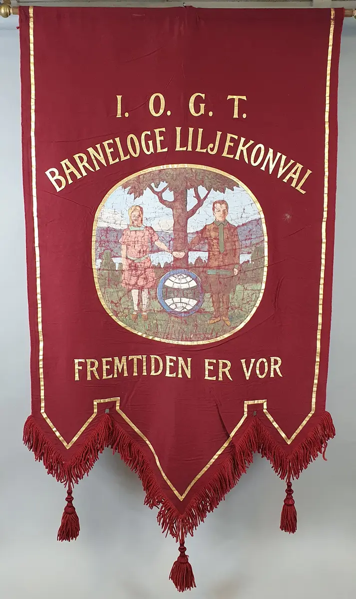 Rød fane med påmalte bokstaver i gull og et malt bilde av to barn - en jente og en gutt som holder hverandre i hendene, og et tre, et vann og fjell i bakgrunnen.
