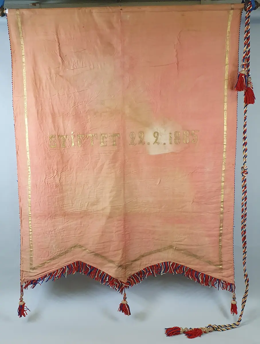 Rød fane i bomull, som er sterkt falmet. Påmalte bokstaver i gull, og et skjoldformet merke med riksløve på svart bakgrunn. Frynser, snor og dusker i rødt, hvitt og blått.