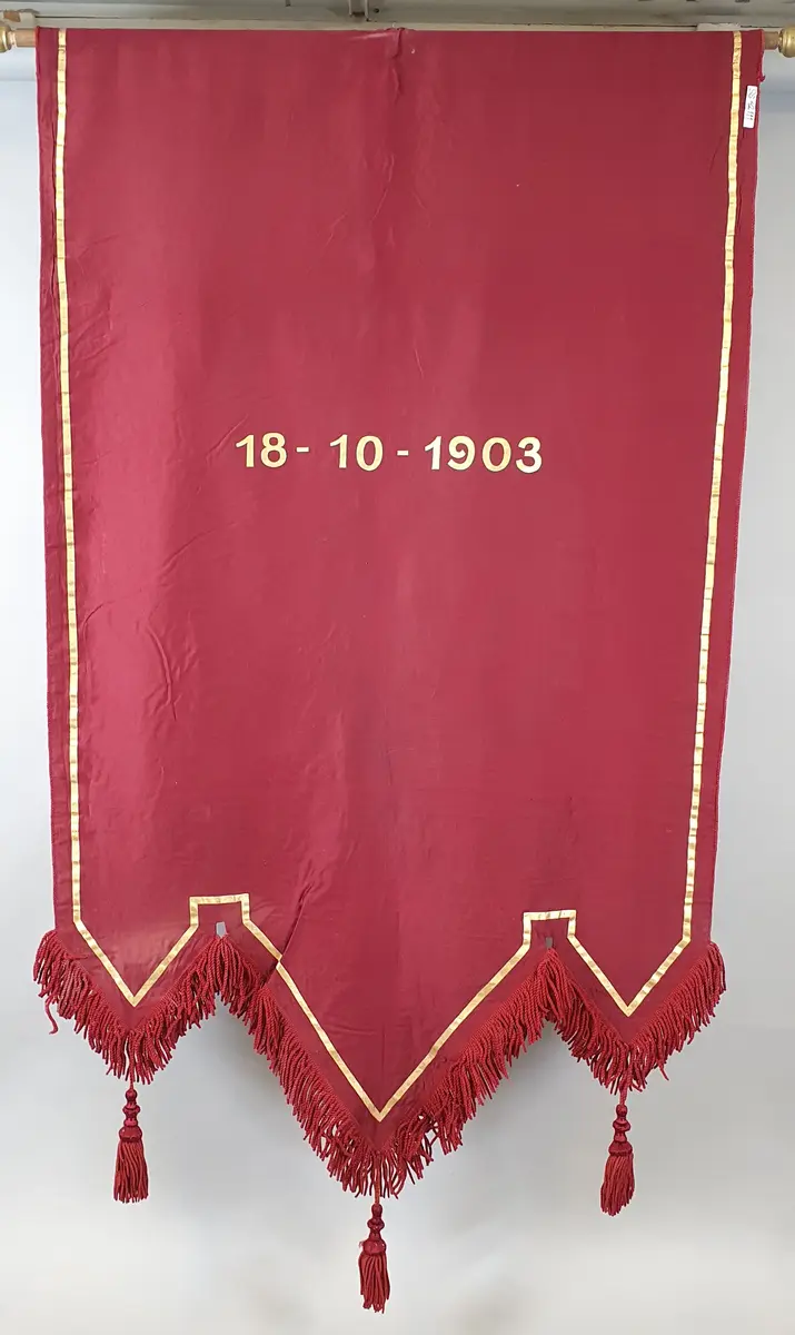 Rød fane med påmalte bokstaver i gull og et malt bilde av to barn - en jente og en gutt som holder hverandre i hendene, og et tre, et vann og fjell i bakgrunnen.