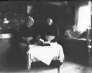 Två okända äldre kvinnor i en stuga. De sitter vid ett lågt bord där en bok ligger uppslagen.