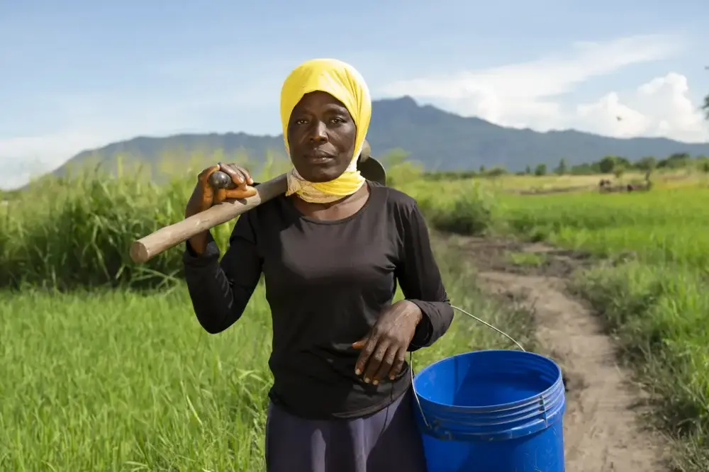 Kvinne. Afrikansk. Med landbruksutstyr over skulderen . Blå bøtte i venstre arm. 