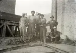 Seks menn i arbeidsklede foran bygningar av murstein. Mogleg