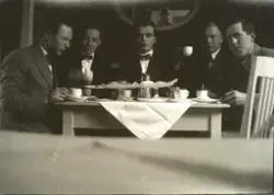 Fem menn ved eit bord dekt med kaffiservise og stettfat.