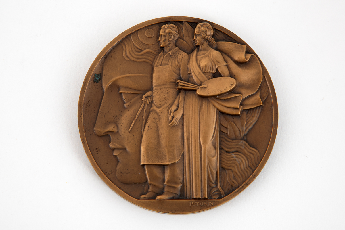 Preget medalje i lavt relieff på begge sider. På avers side et kvinnehode i profil mot venstre med kokarde og laurbærkrans. Mot dette et stående par, vendt mot venstre. Mannen med passer og kvinnen med palett og pensler. Signatur i avskjæringen. På revers side komposisjon med Merkurstav, 2 fakler, hammer og laurbærgren. Omskrift.