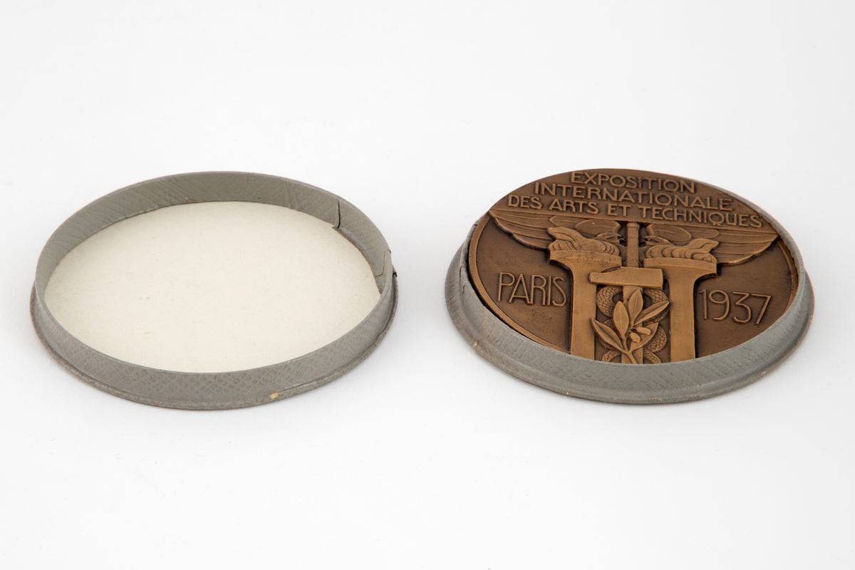 Preget medalje i lavt relieff på begge sider. På avers side et kvinnehode i profil mot venstre med kokarde og laurbærkrans. Mot dette et stående par, vendt mot venstre. Mannen med passer og kvinnen med palett og pensler. Signatur i avskjæringen. På revers side komposisjon med Merkurstav, 2 fakler, hammer og laurbærgren. Omskrift.