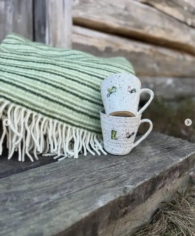 En grön ullpläd och två keramikmuggar står på trappan till ett grånat timmerhus på friluftsmuseet