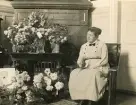 Interiör inifrån borgmästarvillan i Sölvesborg, som idag huserar Hotell Humbla, uppfördes 1903 av borgmästare Philibert Humbla (1863-1925). Far till Philibert Humbla, museiman och svensk konsthistoriker (1896-1952) som 1940 blev förste chef vid Gävle museum (källa Wikipedia). En kvinna sitter framför en kakelugn med ett stor antal blombuketter, troligen en bemärkelsedag.