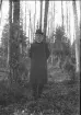 Porträtt av okänd med i en skog, klädd i rock och med kubb på huvudet.