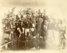 Gruppbild av besättningen utklädda för linjedop på ångkorvetten SAGAs däck i Montevideo 1888-1889. Bland utklädslarna förekommer lösskägg och -hår, klänningar, kavajer, målade dräkter, kronor och hattar. I hänterna håller de bland annat en bok, jättesaxar, ett svärd, blommor, megafon och en treudd. Flera av männen har målat sina kroppar helt svarta i blackfacesmikning.