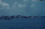 Segelbåtar ligger för ankar i en hamn. I bakgrunden syns en låg kuststräcka med bostadshus.