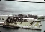 Hotellet Bermudiana i Hamilton, Bermuda sett från vattnet. I förgrunden syns en park omgiven av en kaj. Till vänster i bild ligger ett mindre passagerarfartyg förtöjt vid kajen. Vid kajens spets har en grupp människor har samlats.