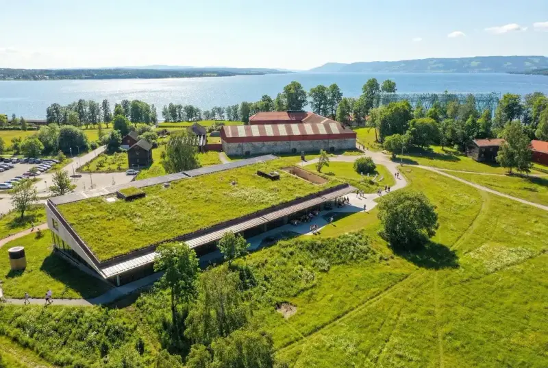 Dronefoto av Anno Domkirkeodden historiesenter med grønt tak, omgitt av grønne trær og kulturlandskap ved Mjøsa.