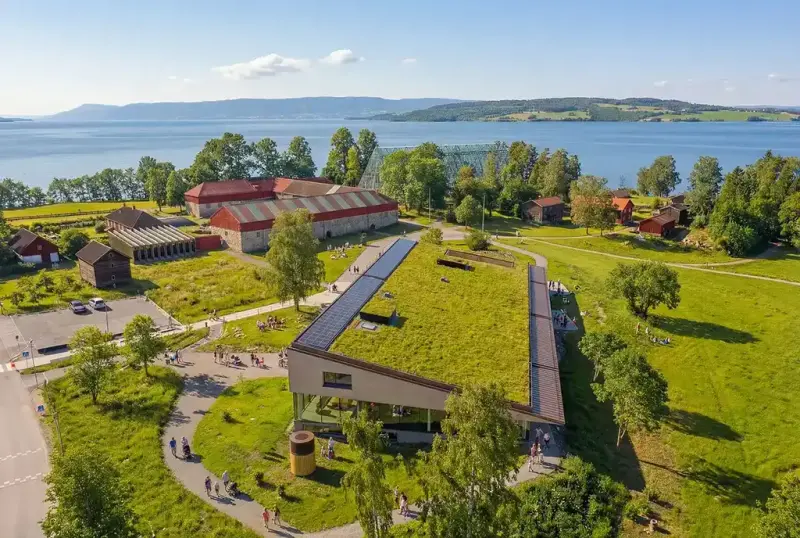 Dronefoto av Anno Domkirkeodden historiesenter med grønt tak, omgitt av grønne trær og kulturlandskap ved Mjøsa.