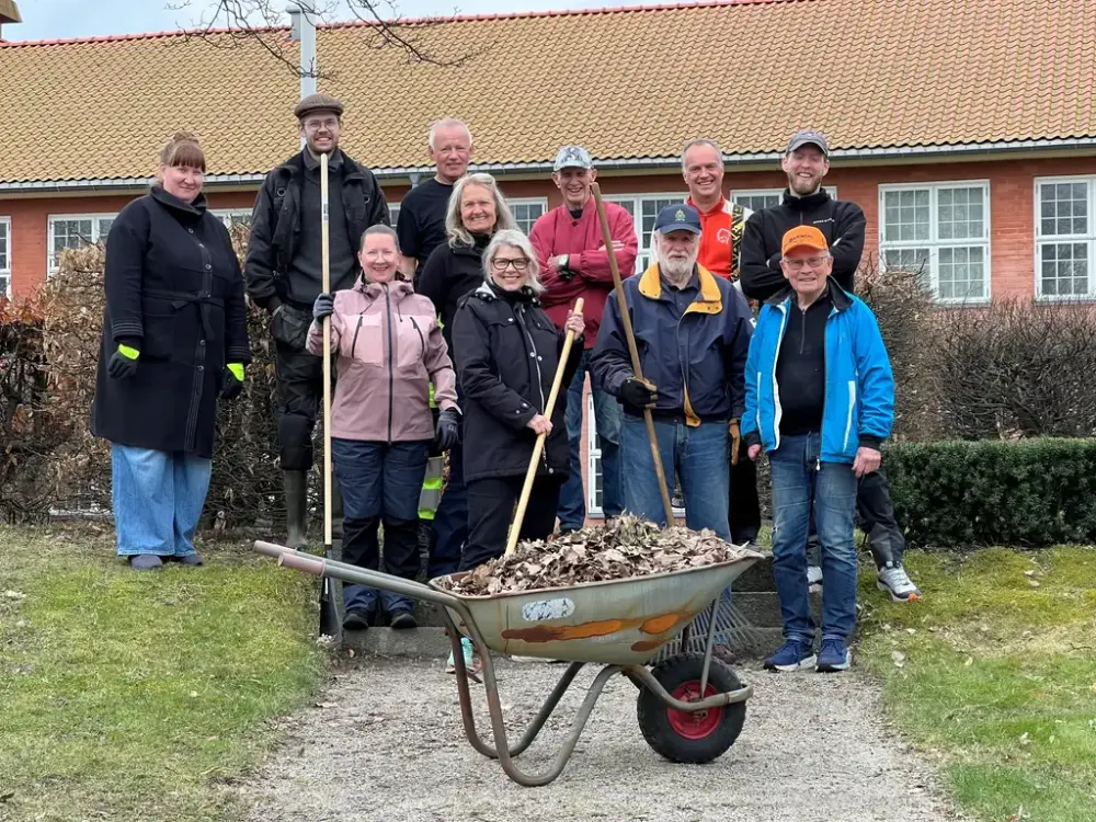 Gruppefoto av en ivrig dugnadsgjeng, bestående av 10 personer, med river og trillebår i forgrunnen