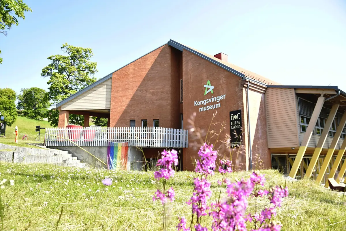 Murbygning med gressplen og villblomster foran og blå himmel bak.