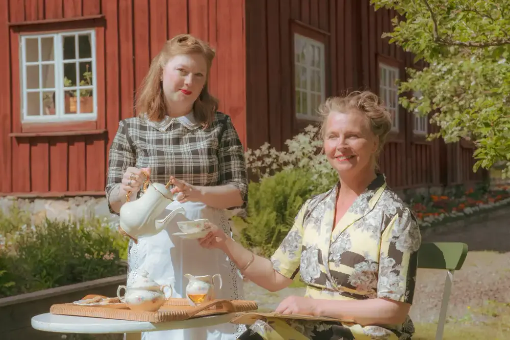 En kvinna serverar en annan sittande kvinna med kaffe framför ett rött hus.