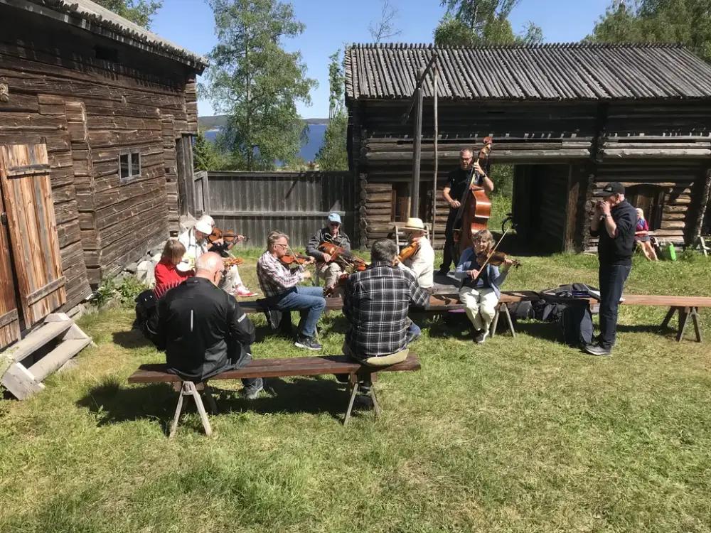 Flera spelmän med olika instrument sitter på bänkar eller står med äldre timmerhus i bakgrunden.