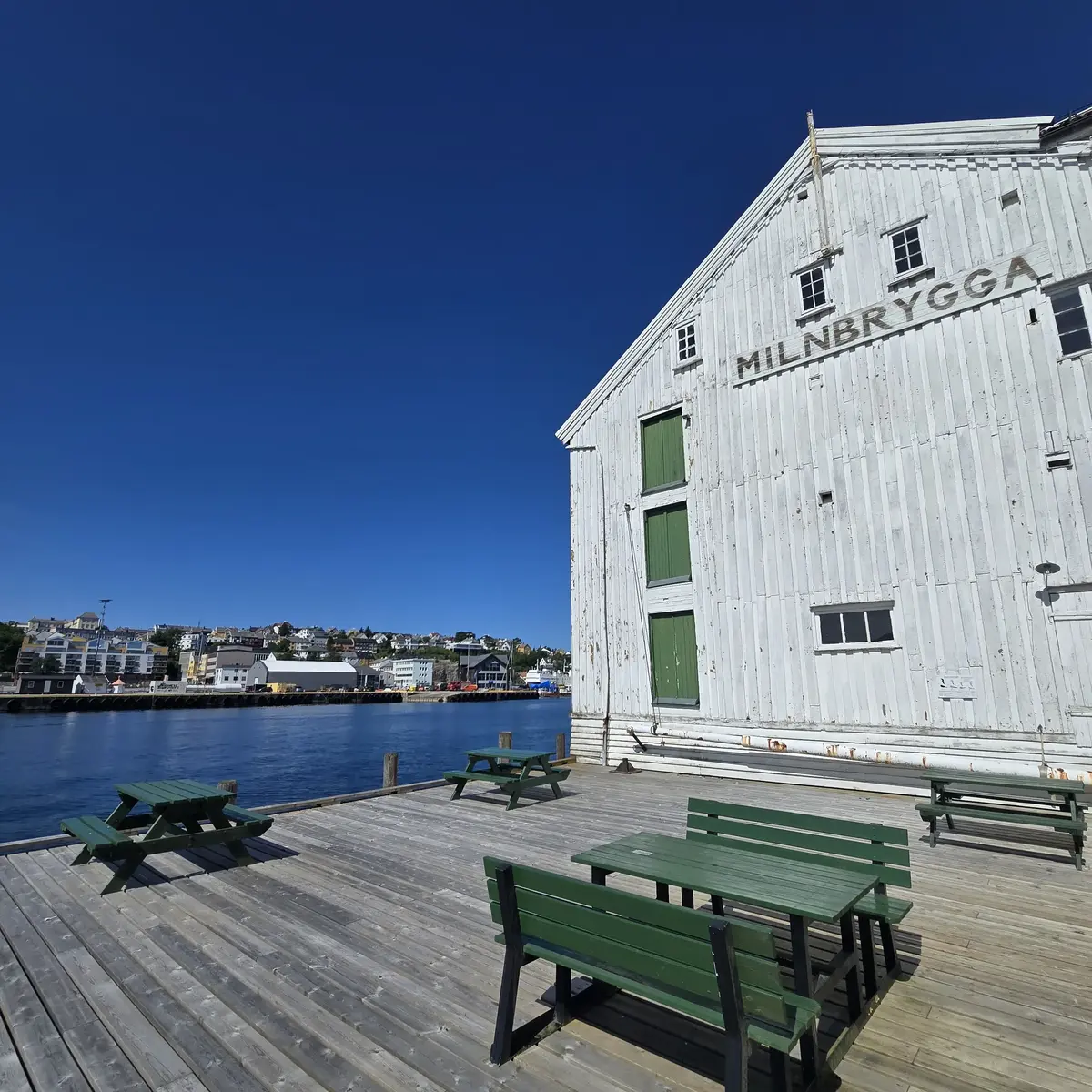 Milnerbrygga som er et gammel trebygg på kaia i Kristiansund, i solskinn, med benker foran.
