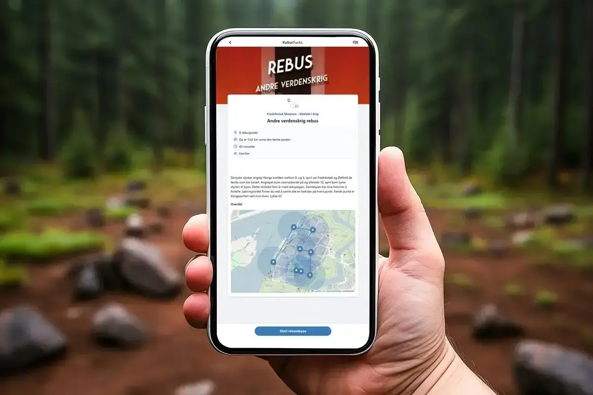 En hånd holder en smarttelefon mot ufokusert skog-bakgrunn. På skjermen vises en digital oppgaveløype ved navn Rebus om 2. verdenskrig