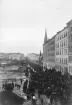 Arbetarrörelsens högtidsdag manifesteras längs Odengatan i Stockholm något av 1900-talets första år. Ett par av huvudpunkterna torde varit krav på rösträtt och bättre arbetsvillkor. På bilden passerar tåget kvarteret Trasten, som i väster pryds av S:t Georgios katedral. Vid den motsatta sidan ligger ännu de blivande kvarteren Lövsångaren och Flugsnapparen obebyggda.