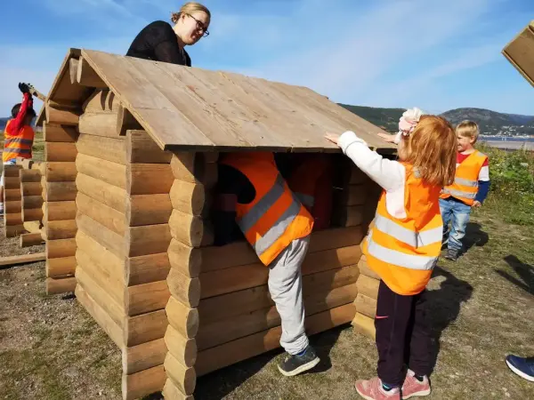 Miniatyr laftehus som bygges av flere barn i oransje vester med 1 voksen. Foto.