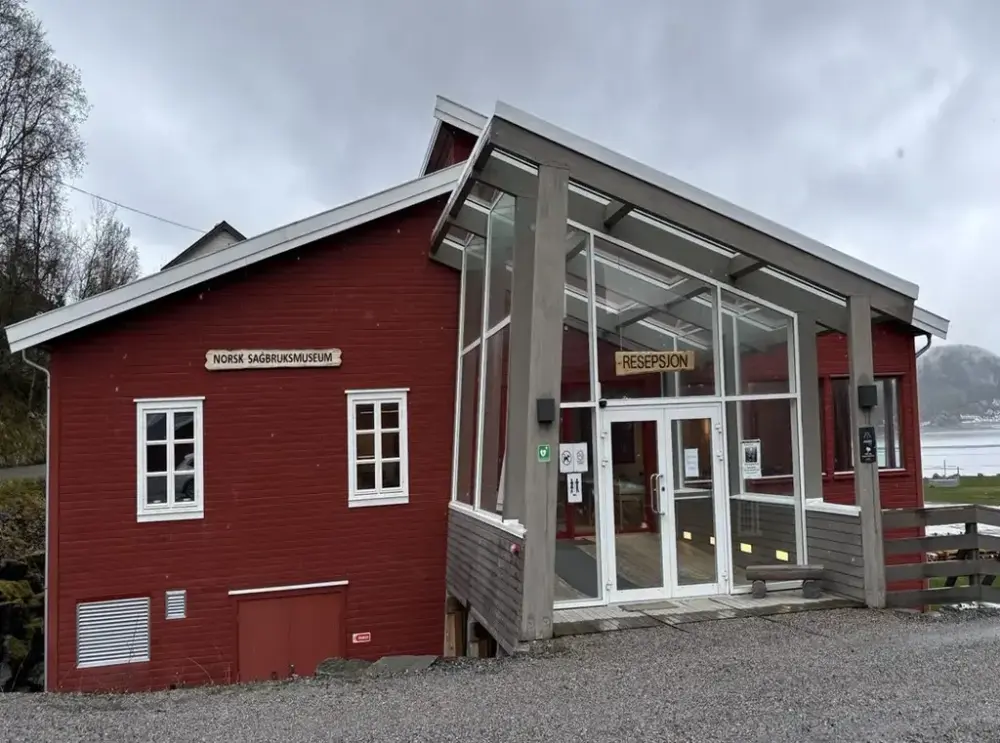 Ombygget rødt trebygg md glassfasade og 2 skilt med Norsk Sagbruksmuseum og Resepsjon. Foto.